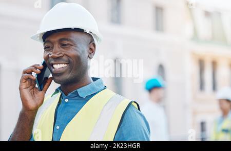 Lavoratori edili che utilizzano uno smartphone per le chiamate in loco. Ritratto di uomo d'affari felice, sorridente e nero in casco di sicurezza e abbigliamento di sicurezza Foto Stock