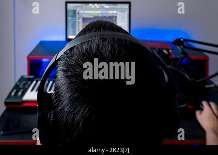 Vista dall'alto posteriore grandangolare dell'uomo con cuffie e capelli neri corti, guardando il monitor e con una mano sul mouse, nello studio di musica di casa, Foto Stock