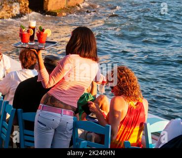 Ripresa da dietro di una cameriera che serve bevande esotiche ai turisti seduti in un bar sul lungomare a Mykonos, Grecia Foto Stock