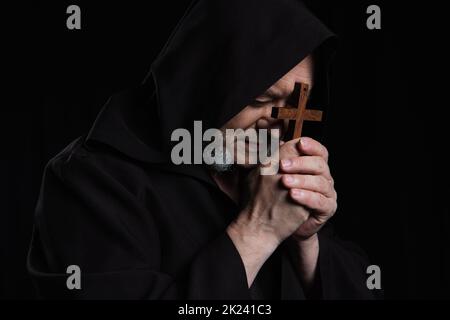 monaco anziano in cassock incappucciati che prega con crocifisso vicino alla faccia isolata su nero, immagine stock Foto Stock