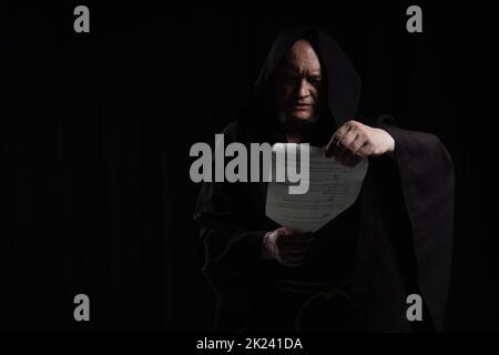 monaco in cassock con cappuccio lettura manoscritto medievale isolato su nero, immagine stock Foto Stock