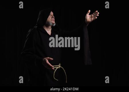 monaco in cassock con cappuccio che punta con la mano mentre prega isolato su nero, immagine stock Foto Stock