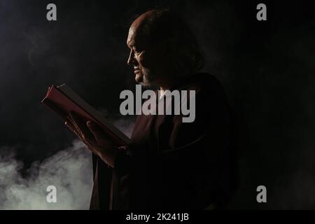 vista laterale del sacerdote anziano in cassock lettura sacra bibbia su nero con fumo, immagine stock Foto Stock