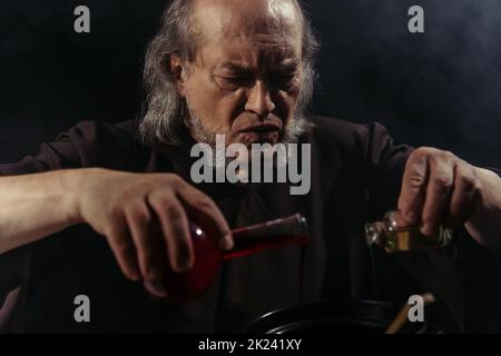 alchimista bearded mescolando ingredienti liquidi e preparando pozione magica isolato su immagine nera, stock Foto Stock