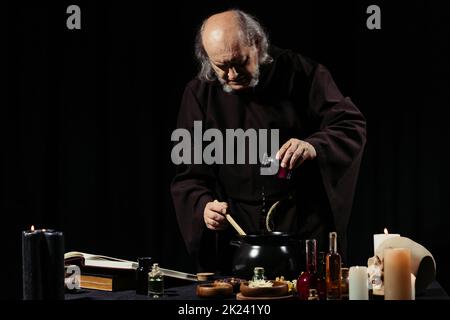 alchimista medievale preparare pozione magica, mentre l'aggiunta di ingrediente in pentola isolato su nero, immagine stock Foto Stock