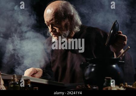 alchimista medievale lettura libro di cucina magica mentre preparando pozione di notte su sfondo nero con fumo, immagine stock Foto Stock