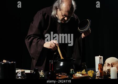 alchimista in vestiario nero preparazione pozione con ingredienti a base di erbe e liquidi isolato su nero, immagine stock Foto Stock