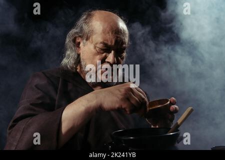 alchimista bearded che aggiunge l'ingrediente nella pentola mentre prepara la pozione su sfondo nero con fumo, immagine di scorta Foto Stock