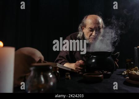 alto alchimista vicino bollitura e pentola di cottura a vapore preparazione pozione di notte isolato su nero, immagine stock Foto Stock