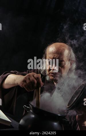 guaritore medievale preparare pozione magica in pentola bollente isolato su nero, immagine stock Foto Stock