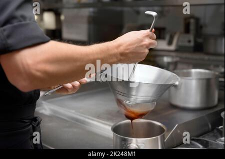 Cucina nella cucina del ristorante. Setacciare la salsa attraverso un setaccio. Lo chef sta filtrando la salsa. Foto di alta qualità Foto Stock