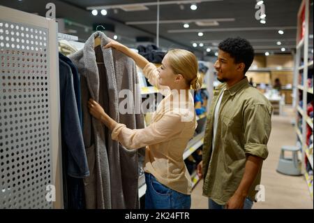 Interracial famiglia coppia cliente scegliere accappatoio in negozio. Donna che acquista vestiti per il marito Foto Stock