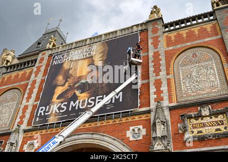 Amsterdam, Paesi Bassi - Agosto 2022: Persone in piedi su una piattaforma di raccolta ciliegie che fissa un grande banner alla parete esterna del famoso Museo Rijks Foto Stock