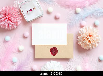 Carta e busta vicino a fiori pastello, pom-pom, piume e anello in una confezione regalo su rosa Foto Stock