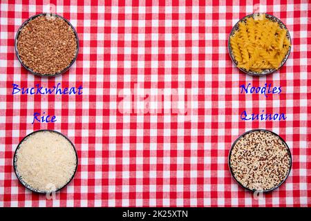 Cibo vegetale. Primo piano di quattro ciotole con grano saraceno, tagliatelle, riso e quinoa su una tovaglia rossa a scacchi isolata. Alimentazione sana. Vista dall'alto con spazio. Foto Stock