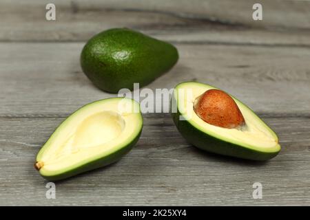 Due avocadi, uno tagliato a metà, seme visibile, sul legno grigio scrivania Foto Stock