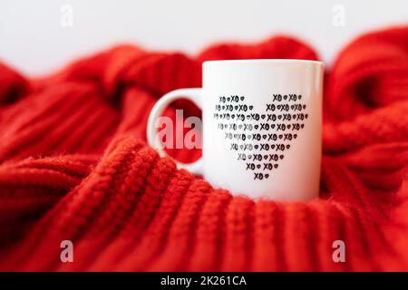 Una tazza bianca con un cuore è in piedi su un tessuto a maglia rosso. Il concetto di amore e celebrazione di San San Valentino, 14th febbraio. Foto Stock