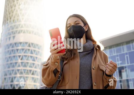 Ritratto di giovane donna d'affari che indossa maschera protettiva FFP2 KN95 camminare in strada moderna città utilizzando il suo telefono Foto Stock