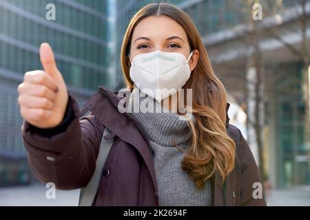 Primo piano di ottimista donna d'affari che indossa maschera protettiva KN95 FFP2 mostrando i pollici in su nella strada moderna della città e guardando la macchina fotografica Foto Stock
