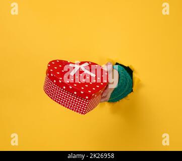 la mano femminile tiene una scatola rossa con un regalo su uno sfondo giallo, parte del corpo sporge da un foro strappato su uno sfondo di carta. Congratulazioni, vacanza sorpresa Foto Stock