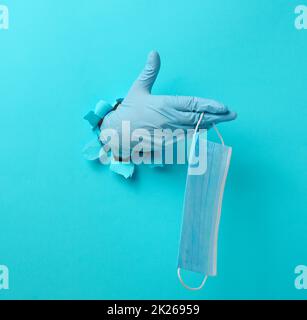 una mano femminile tiene una maschera medica usa e getta per proteggere contro i virus durante un'epidemia e pandemia. Parte del corpo sporge da un foro strappato in carta blu Foto Stock