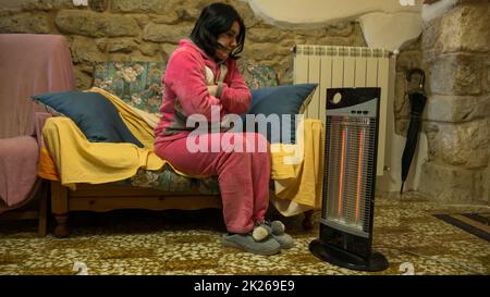 bella donna fredda in pigiama rosa che si trova sul divano a casa scaldando le mani e i piedi di fronte ad un riscaldatore elettrico. brivido nel calore di casa. Foto Stock