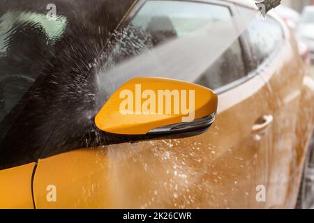 Lo specchietto laterale di giallo auto lavata in self service autolavaggi, spruzzo a getto di acqua che colpisce la finestra di gocce di acqua volare da tutti i lati. Foto Stock