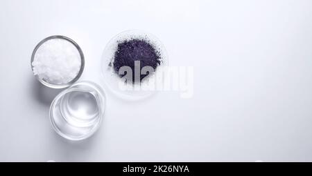 Ingrediente chimico cosmetico su tavolo bianco da laboratorio (vista dall'alto). Cera microcristallina in contenitore di vetro, KMnO4 in vetro da orologio chimico e alcool in becher. Foto Stock