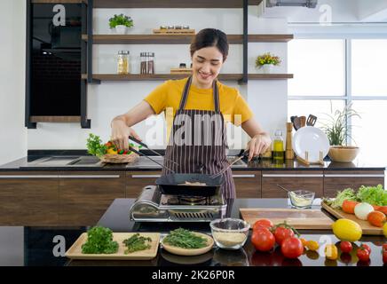 Giovane casalinga che usa BBQ Clamp Shovel e acciaio inossidabile forcella tostare la bistecca. Atmosfera mattutina in una cucina moderna. Il banco della cucina è pieno di vari tipi di verdure e condimenti. Foto Stock