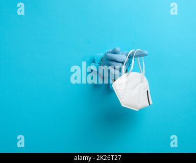 la mano femminile tiene una maschera medica monouso bianca per proteggere contro i virus durante un'epidemia e pandemia. Parte del corpo sporge da un foro strappato in carta blu Foto Stock