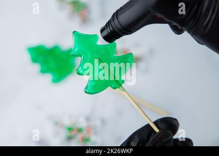 Un lecca lecca verde sotto forma di albero di Natale è tenuto nelle mani di guanti neri e bruciato sul fuoco da un accendino, nel processo di fabbricazione di un lecca. Foto Stock