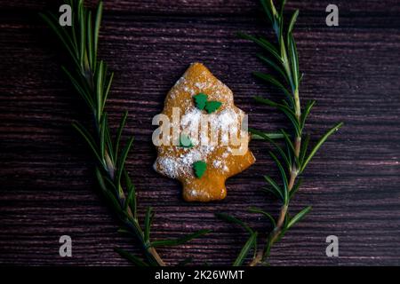 Biscotti sotto forma di albero di Natale, cosparsi di zucchero a velo e piccoli alberi di Natale in cima, si trova su uno sfondo di legno scuro circondato da rosmarino granato Foto Stock