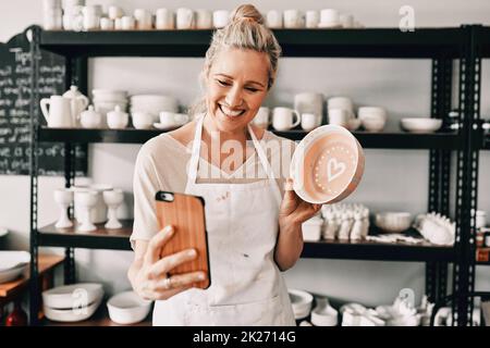 Ehi, guarda ragazzi. Scatto corto di una donna matura attraente usando il suo cellulare per prendere un selfie con la sua ceramica nel suo studio. Foto Stock