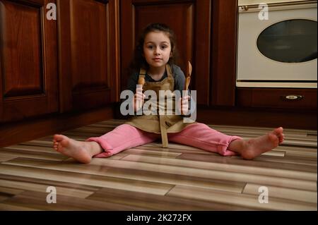 Ragazza cute del bambino, pasticciere piccolo del panettiere nel grembiule dello chef gioca con i cucchiai di legno, seduta a piedi nudi sul pavimento della cucina Foto Stock