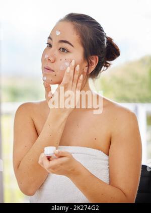 Mantenere la mia faccia idratata tutto il giorno. Shot di una giovane donna che applica moisturizer al suo viso durante la sua routine di bellezza mattutina. Foto Stock