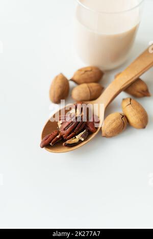 Latte di pecan vegano in una tazza di vetro con noci intere su un tavolo bianco, noci in un cucchiaio di legno. Foto Stock