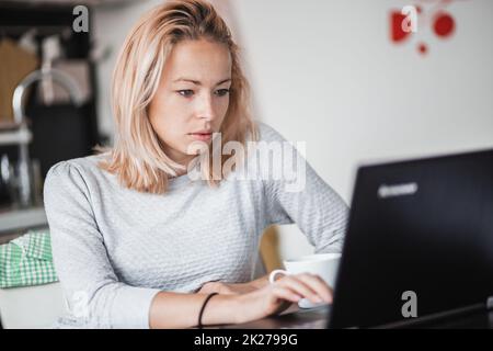 Freelance femmina nel suo casual home abbigliamento lavorando in remoto dal suo tavolo da pranzo al mattino. Cucina di casa in background. Foto Stock