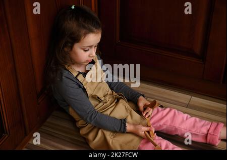 Adorabile bambina in grembiule beige dello chef, giocando con utensili da cucina in legno, seduta a piedi nudi sul pavimento Foto Stock