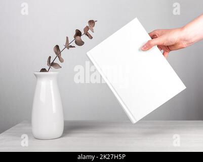 Mano che tiene il libro mockup vicino ai fiori in vaso a tavola. Stile minimalista. Lettura del tempo libero, concetto di armonia. Romanzo con copertina bianca. Foto di alta qualità Foto Stock