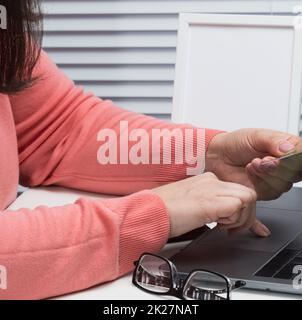 donna in un maglione rosa effettua acquisti online con un laptop, una carta di credito in mano. Una donna siede su un tavolo bianco Foto Stock