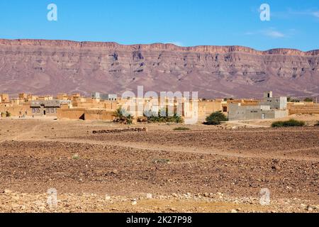 Tipico paesaggio desertico nel sud del marocco, piccolo villaggio con edifici rettangolari in pietra, pochi cespugli verdi, montagne a distanza Foto Stock