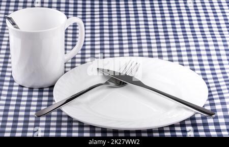 Empy Mug con cucchiaio, piatto con un coltello e forchetta sulla tovaglia in una gabbia. Vista dall'alto con spazio per la copia Foto Stock