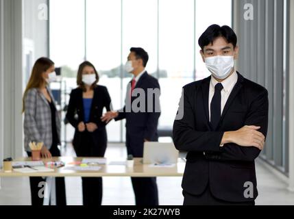 Giovane uomo d'affari asiatico in vestito nero ripiegare le braccia sul petto con fiducia. Il team si sta preparando all'incontro in background. Tutti indossano una maschera facciale. Foto Stock