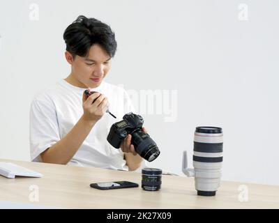 Il giovane fotografo asiatico sta pulendo lo schermo della fotocamera e il corpo con una fotocamera soffiante per la pulizia sulla sua scrivania. Foto Stock