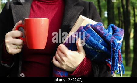 Una tazza di caffè rossa e un libro con una coperta di lana a scacchi blu o un plaid nelle mani di una donna che indossa un maglione e un cappotto nero nel parco. Tempo caldo e soleggiato. Fotografia morbida e accogliente Foto Stock