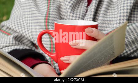 Una ragazza vestita con un cappotto e una sciarpa nella foresta autunnale tiene un libro e una tazza con una bevanda calda nelle sue mani primo piano in un parco cittadino in una giornata calda. Il concetto di lettura, relax e comfort. Foto Stock