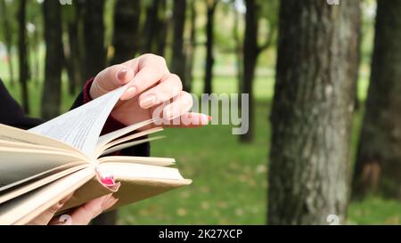 Ragazza che legge un libro nel parco. Le mani femminili sfogliano pagine di libri di carta all'aperto. Lo studente si sta preparando per l'esame. Tempo libero letterario in natura. Primo piano, spazio di copia. Foto Stock