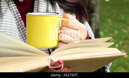 Una ragazza vestita con un cappotto e una sciarpa nella foresta autunnale tiene un libro e una tazza con una bevanda calda nelle sue mani primo piano in un parco cittadino in una giornata calda. Il concetto di lettura, relax e comfort. Foto Stock