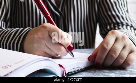 Vista laterale delle mani di una donna con unghie rosse, tenendo una penna rossa, scrive qualcosa su un foglio vuoto di carta da agenda giacente su una tavola grigia. La ragazza prende appunti in un notebook nero Foto Stock