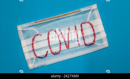 Tipica maschera chirurgica bianca a 3 strati con auricolari in gomma per coprire bocca e naso con lettere a blocco inglesi Covid su sfondo blu. Virus pericoloso e concetto di quarantena. Foto Stock